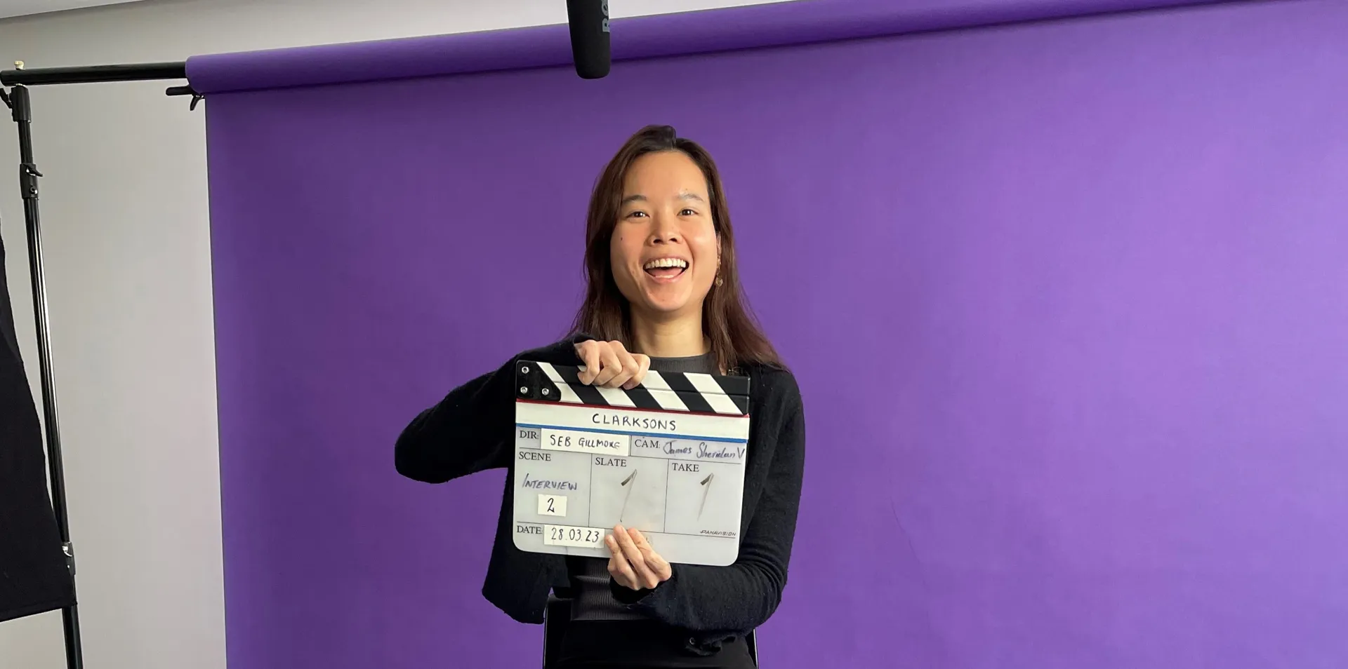 woman sitting on a stool behind a purple background, smiling, holding a clapper